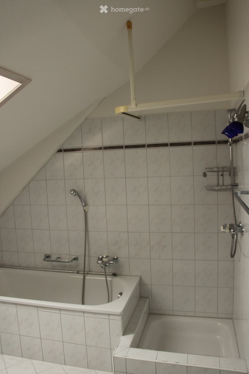 Bathroom with a bathtub, tiled walls, and a showerhead. Features include a handrail next to the bathtub and a window at the top left corner.