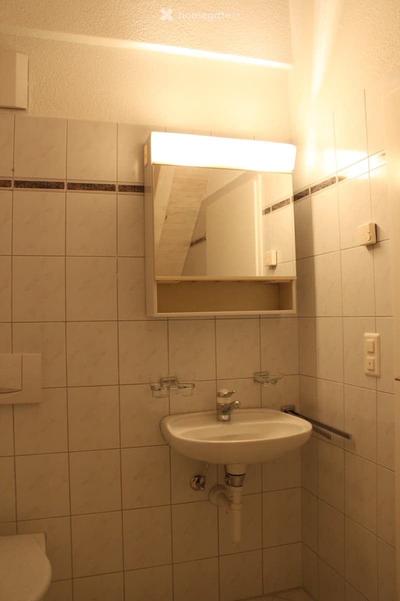 A small bathroom with a white tiled wall, a sink with a chrome faucet, a mirror with built-in lighting, and a cup holder on the wall.