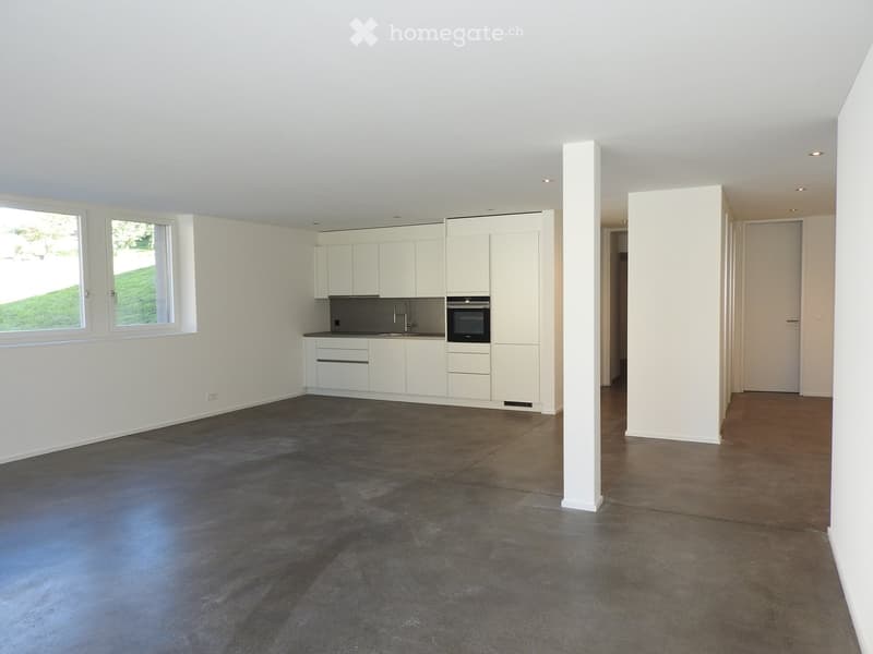 Empty room with white walls, gray floor, kitchen area with cabinets, sink, oven, and window