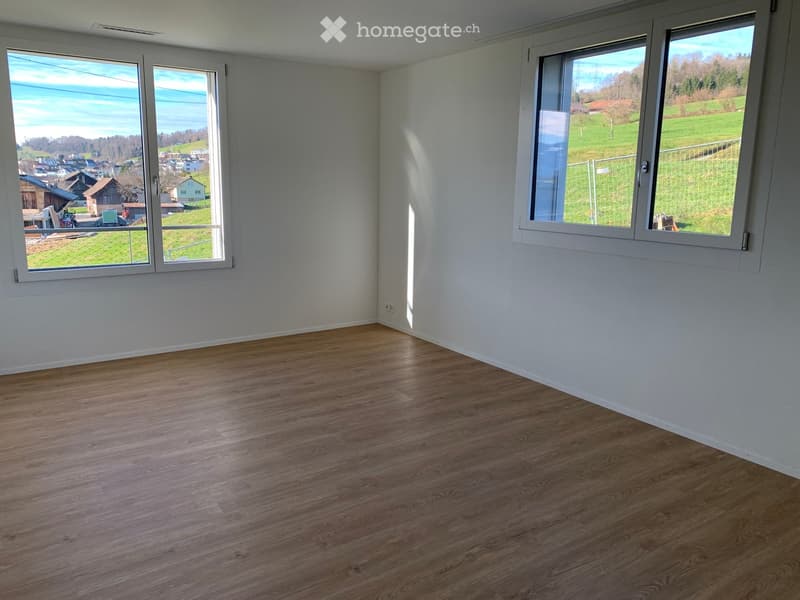 Empty room with wooden flooring and large windows offering a scenic view of a green landscape and distant houses.