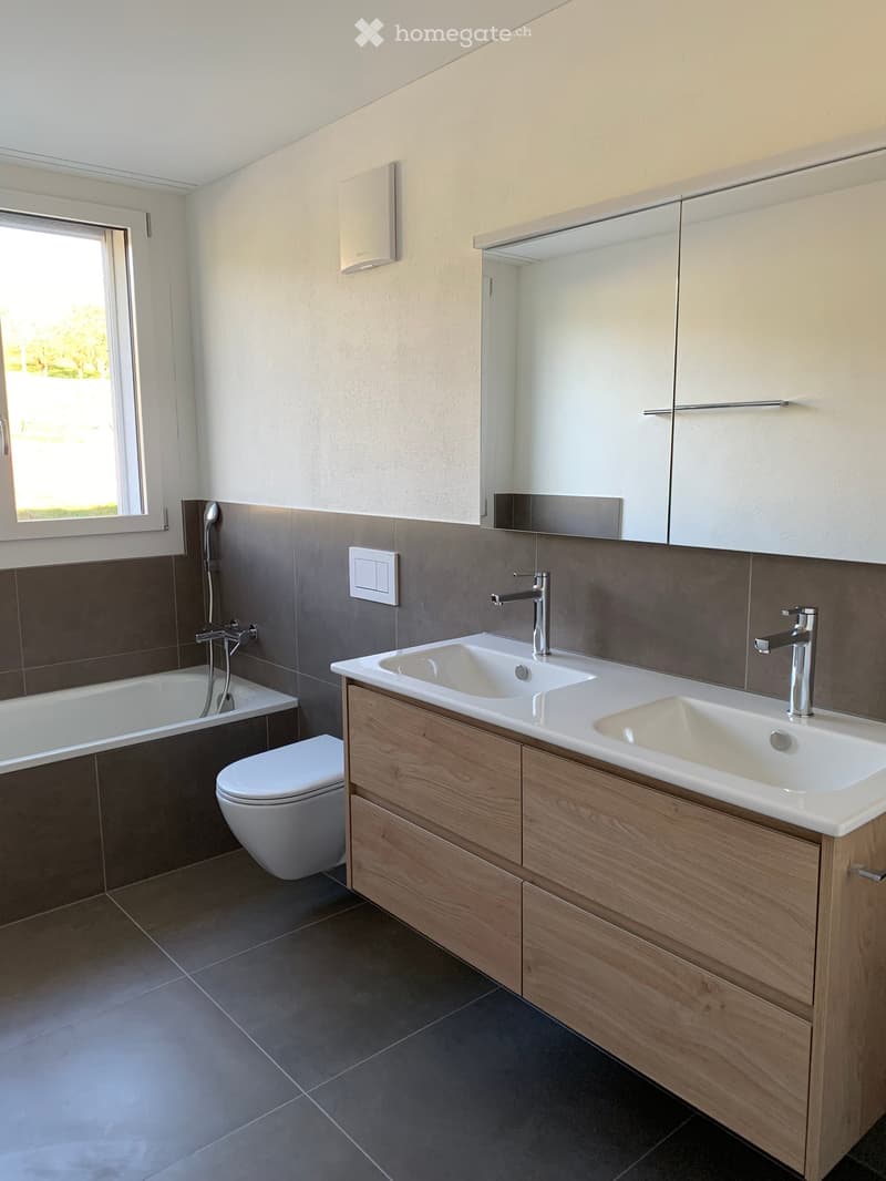 Modern bathroom with a double sink, wooden cabinet, bathtub, toilet, and large mirror. The walls are tiled in dark brown and light beige, and there is a window providing natural light.