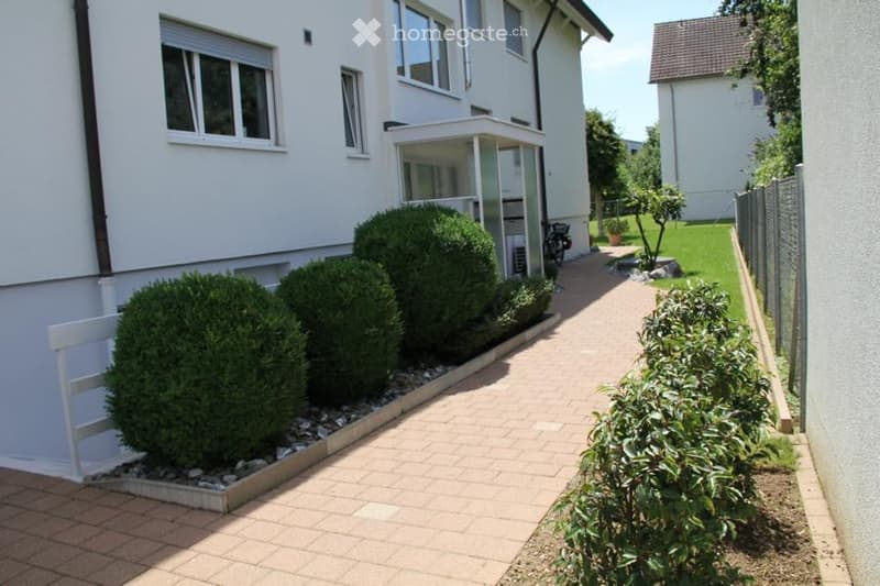 Well-maintained garden area with neatly trimmed bushes, a brick pathway, and a small white bench near the building entrance.