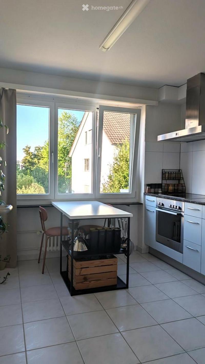 Modern kitchen with white cabinets, stainless steel oven, large windows, and a small table with a chair.