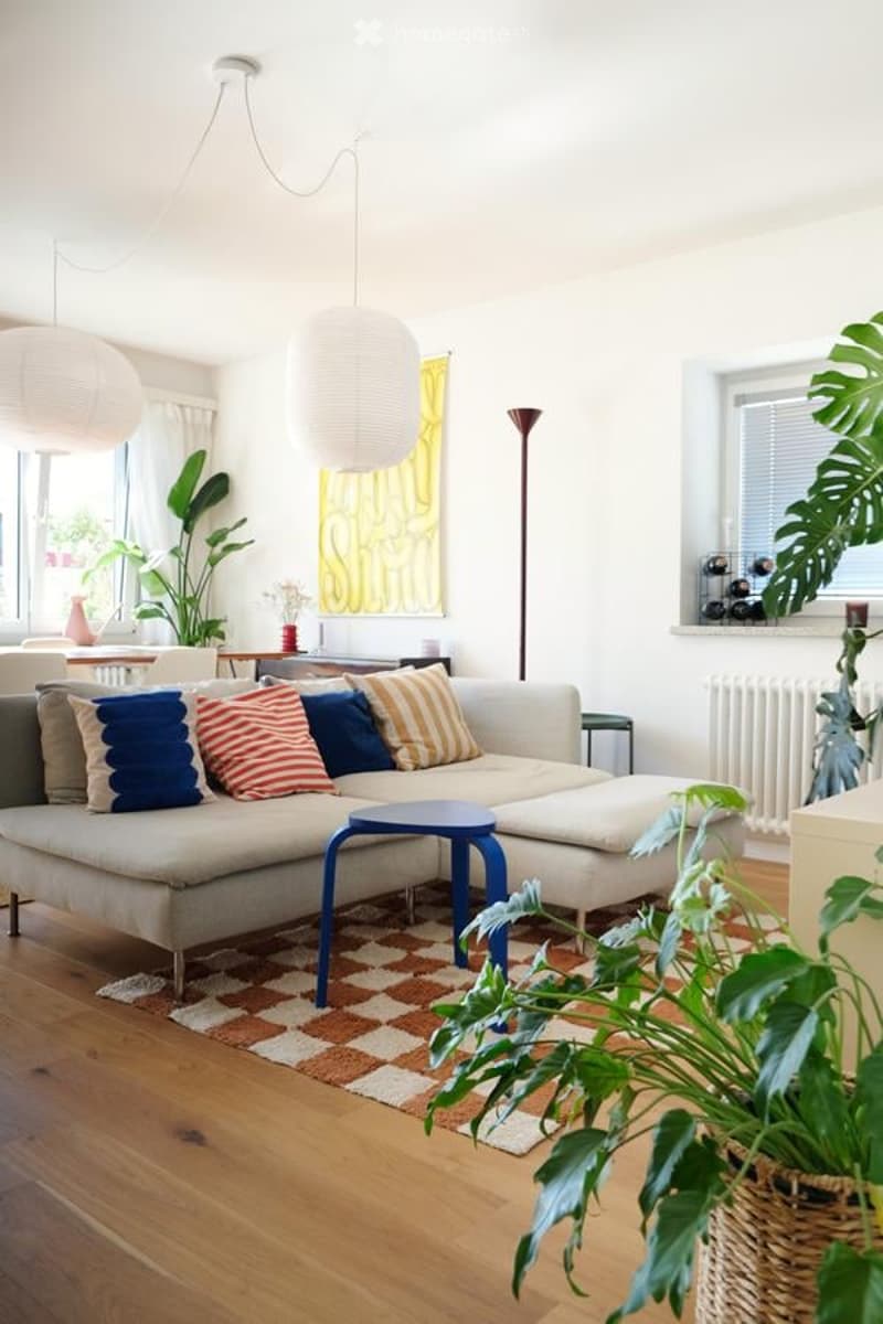 Modern living room with a grey sectional sofa, colorful throw pillows, a blue coffee table, hanging paper lanterns, and potted plants. The room features wooden flooring and large windows with white blinds.
