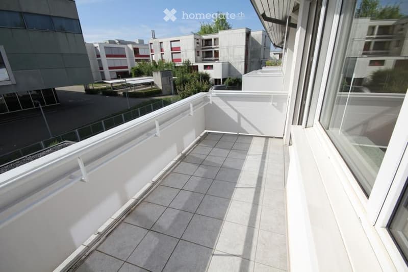Modern balcony with white tiled flooring, white railings, and large windows overlooking a contemporary building complex.