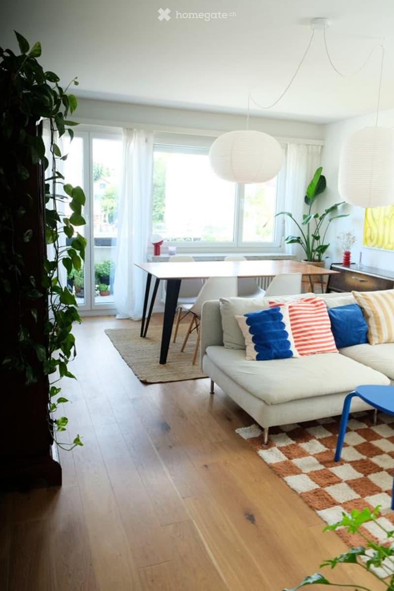 Bright living room with wooden flooring, large windows, and a balcony. Features a grey sectional sofa with colorful throw pillows, a coffee table, a dining table with chairs, pendant lights, and indoor plants.