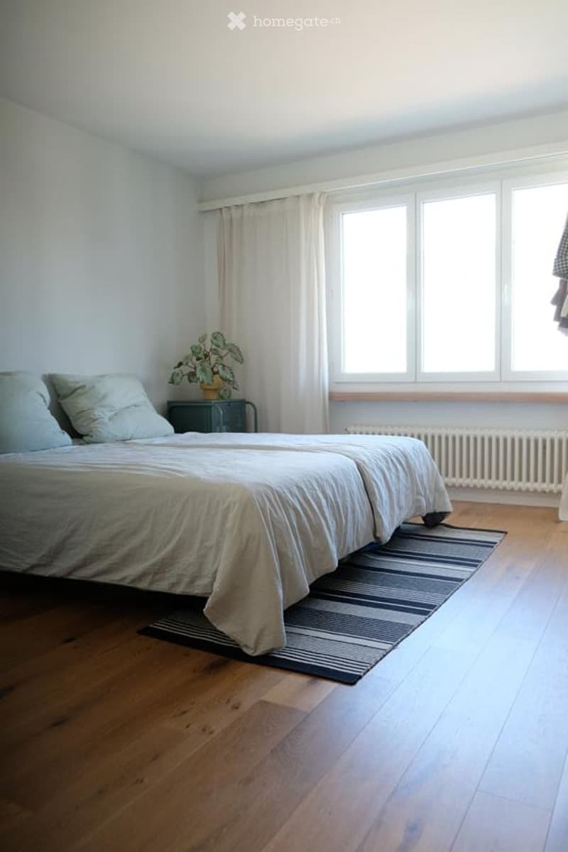 Bright bedroom with wooden flooring, large windows with white curtains, a neatly made bed, a small nightstand with a plant, and a radiator below the windows.