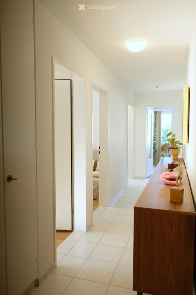 Bright hallway with white tiled flooring, white walls, and recessed lighting. Features open doors leading to adjacent rooms and a small console table with decorative items on the right side.