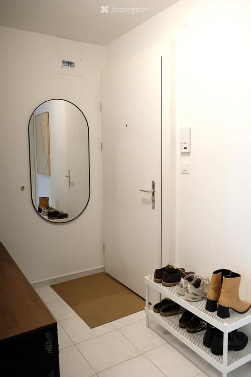 Clean, minimalist hallway with white walls, tiled floor, and a shoe rack holding multiple pairs of shoes. A round mirror on the wall reflects part of the adjacent room.