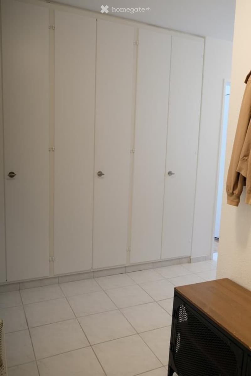 Empty room with white tiled flooring and white built-in wardrobes. A small wooden table and a coat hanging on the wall are visible.