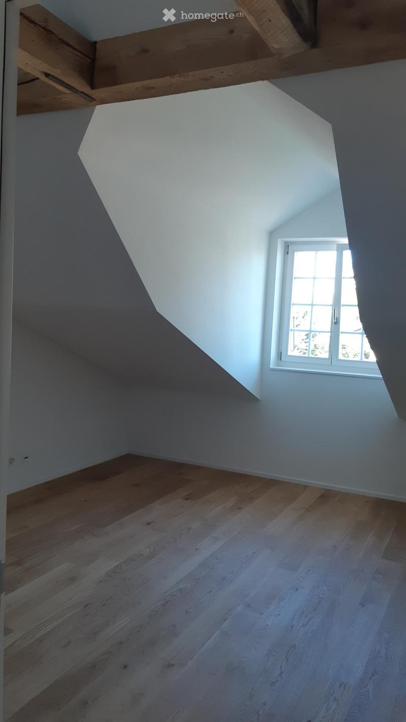 Empty attic space with wooden flooring, white walls, and a single window letting in natural light. Exposed wooden beams on the ceiling.