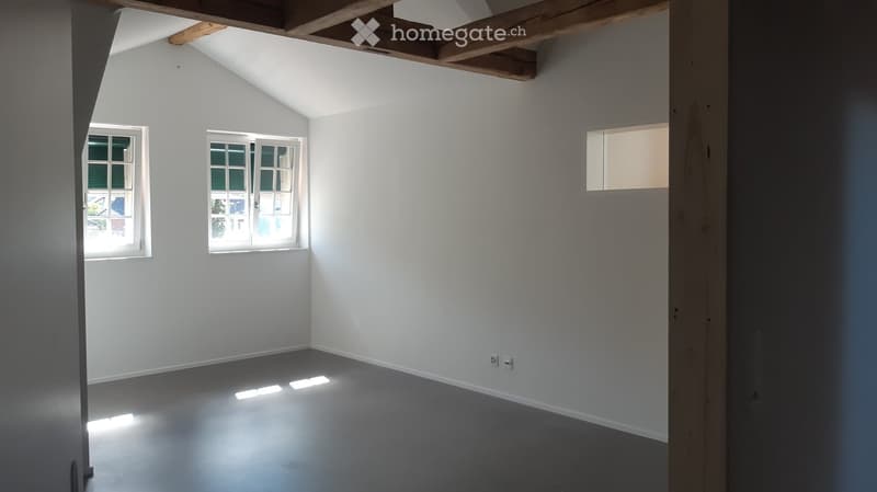 Empty room with white walls, grey floor, and three windows allowing natural light. Exposed wooden beams on the ceiling.