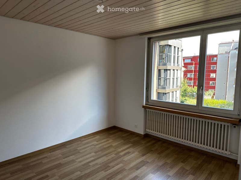 Empty room with wooden flooring, white walls, and a large window with a radiator below it. The room has a slanted wooden ceiling and a view of neighboring buildings.