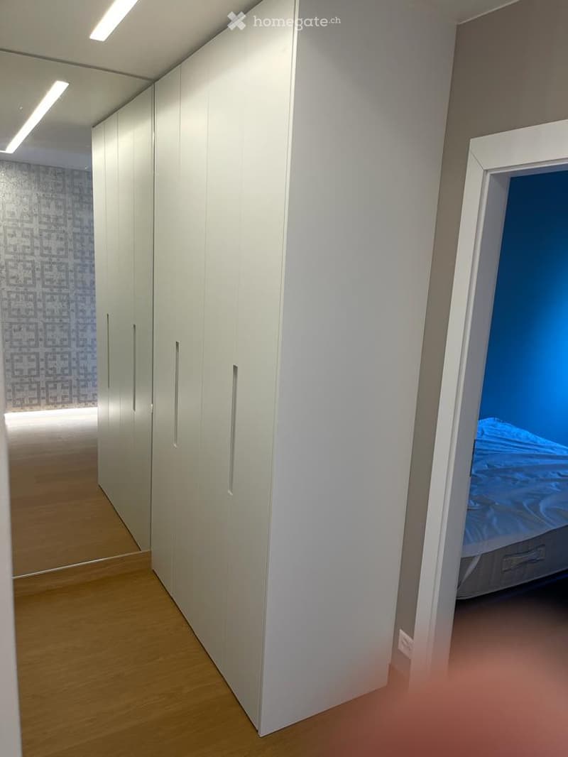 A modern, minimalist hallway with white built-in wardrobes and a glimpse of a blue bed in an adjacent room.
