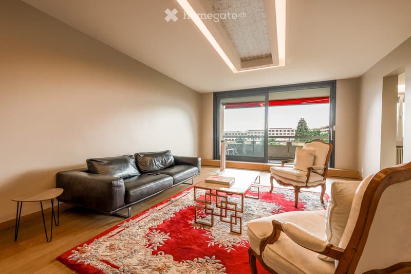Modern living room with a slanted ceiling, large windows, and a balcony. Features a black leather sofa, white armchairs, a coffee table, and a red patterned rug.