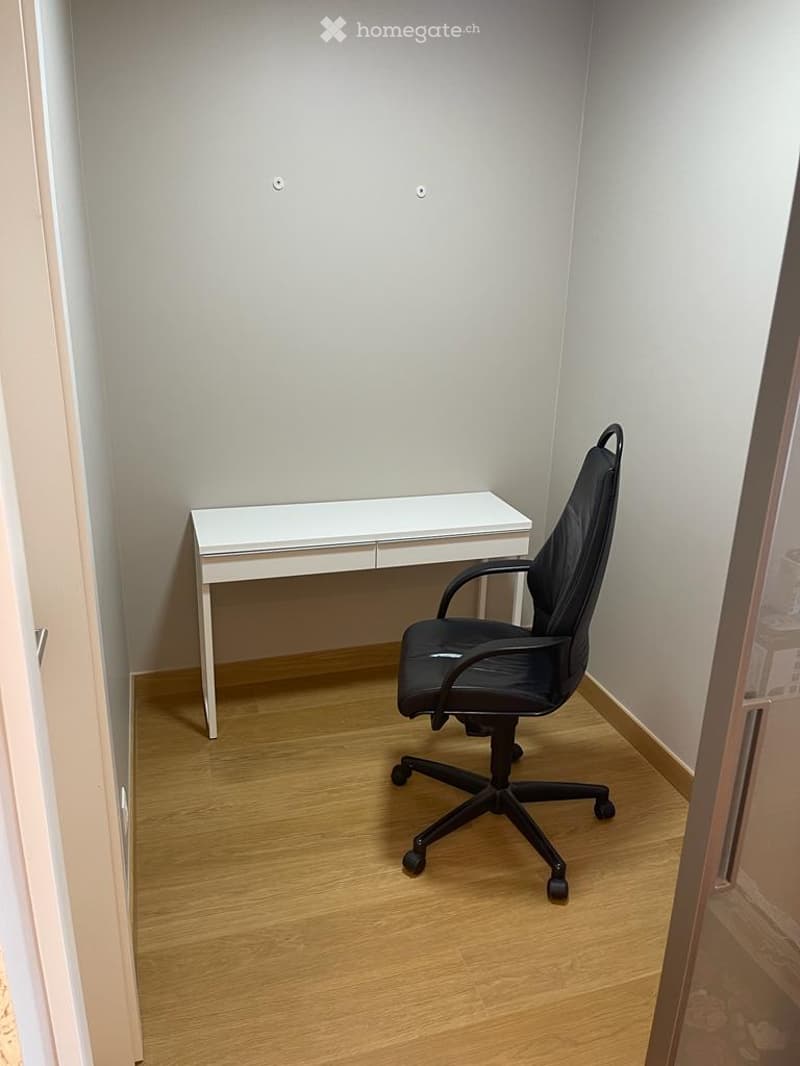A small, unfurnished room with a white desk and a black office chair, wooden flooring, and light grey walls.