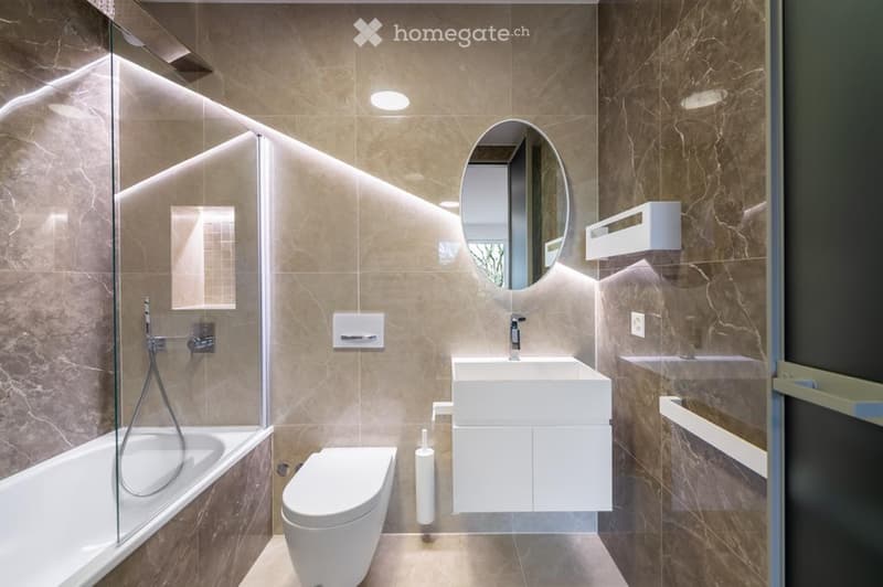 Modern bathroom with marble tiles, a bathtub, toilet, sink, and a large oval mirror above the sink.