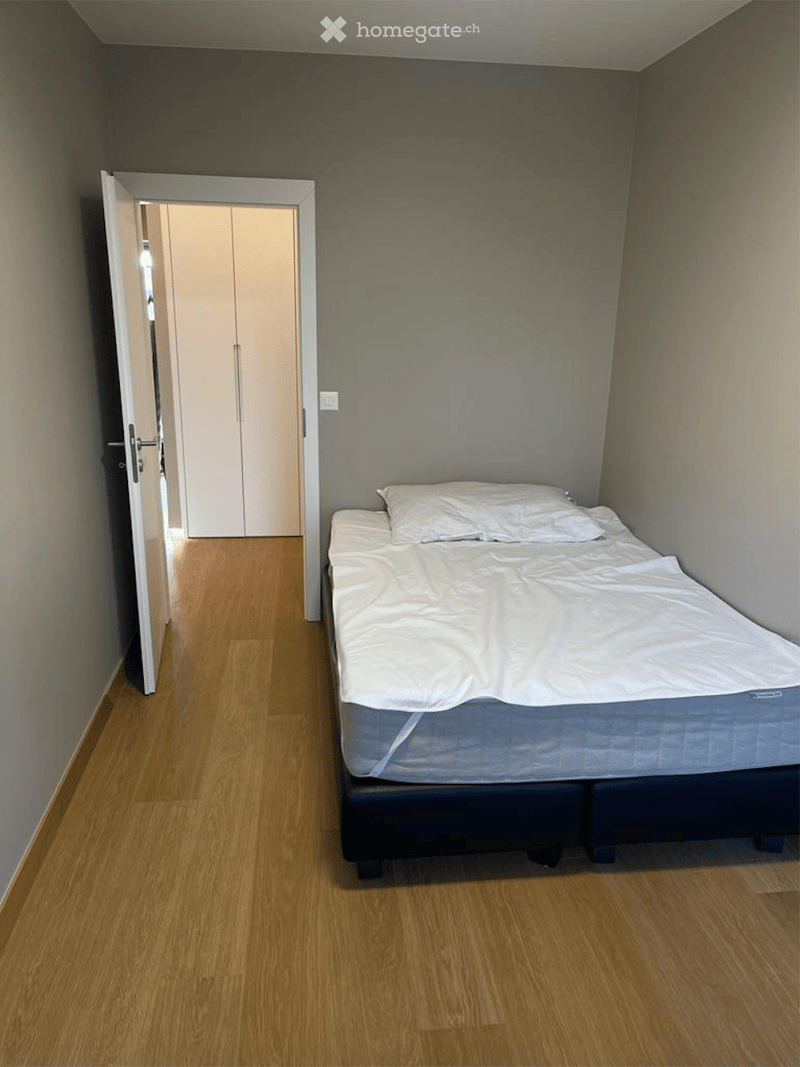 A small, neatly furnished bedroom with a single bed, white bedding, and a wooden floor. An open closet is visible in the background.