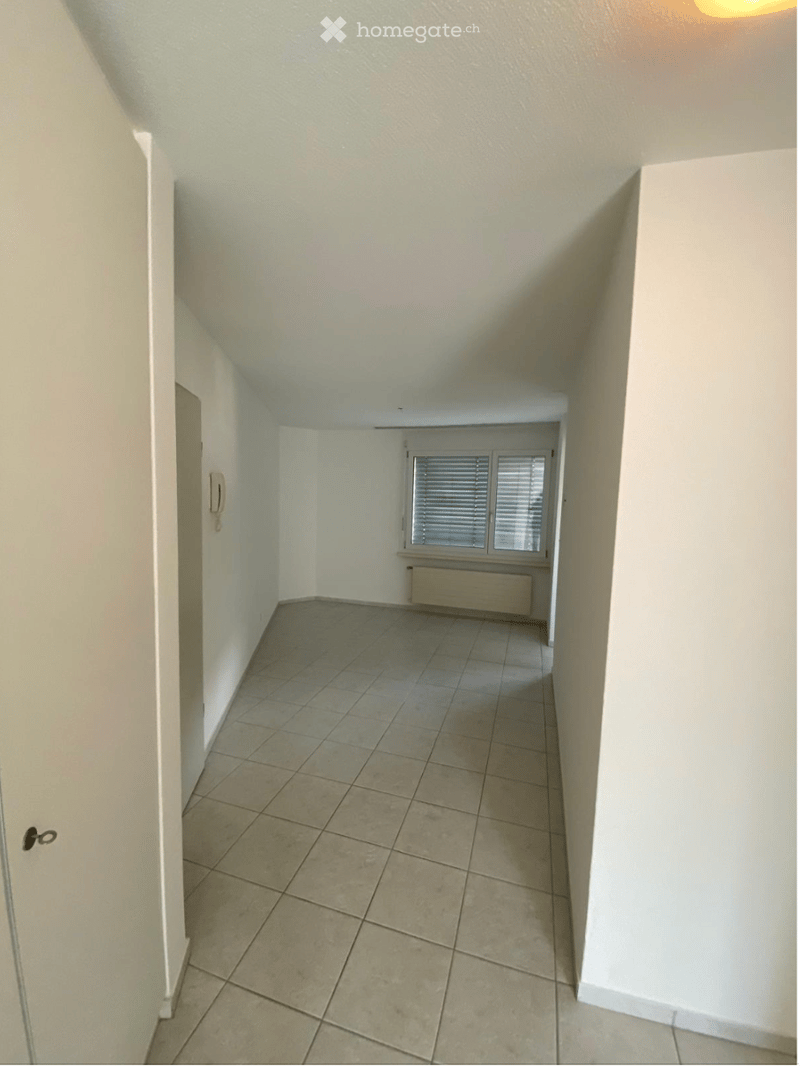 Empty hallway with tiled floor, white walls, and a window with blinds at the end.