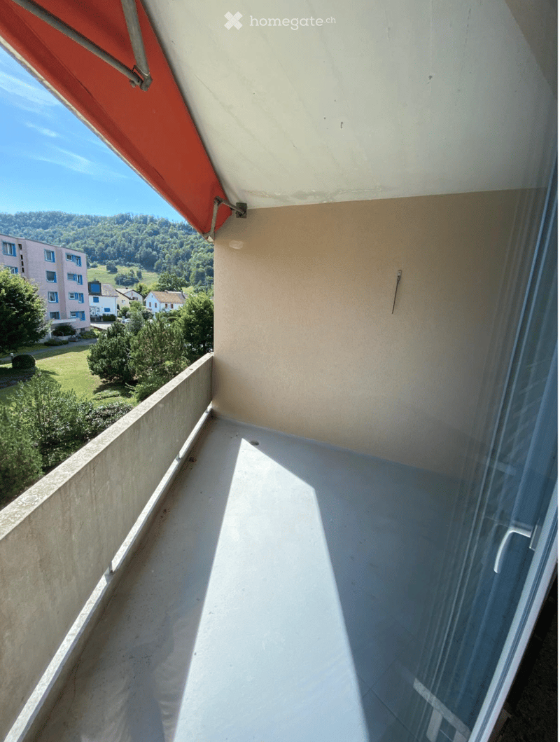 Balcony with a concrete floor, glass railing, and a red awning, overlooking a green landscape with trees and buildings.