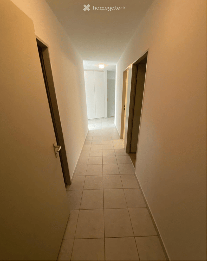 A clean, well-lit hallway with tiled flooring and white walls, featuring several doors and a ceiling light fixture.