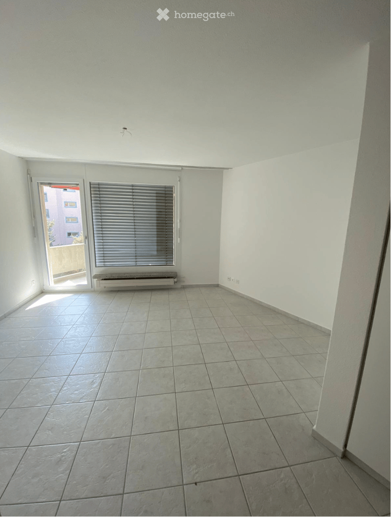 Empty room with tiled floor, white walls, large window with blinds, and a small bench beneath the window.