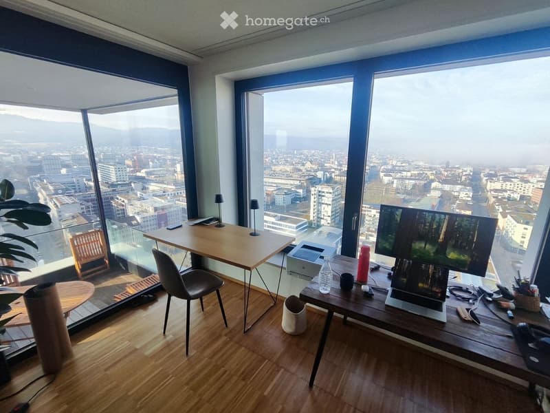 Modern, well-lit room with large windows offering a cityscape view. Features a wooden desk with a laptop, screen, printer, and various office supplies, along with a comfortable chair and additional seating area with a small table and chair.