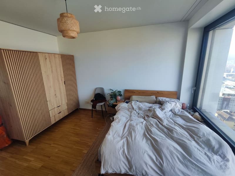 Modern bedroom with wooden floor, large window offering city view, bed with white bedding, wooden wardrobe, nightstand with plant, and hanging wicker light fixture.
