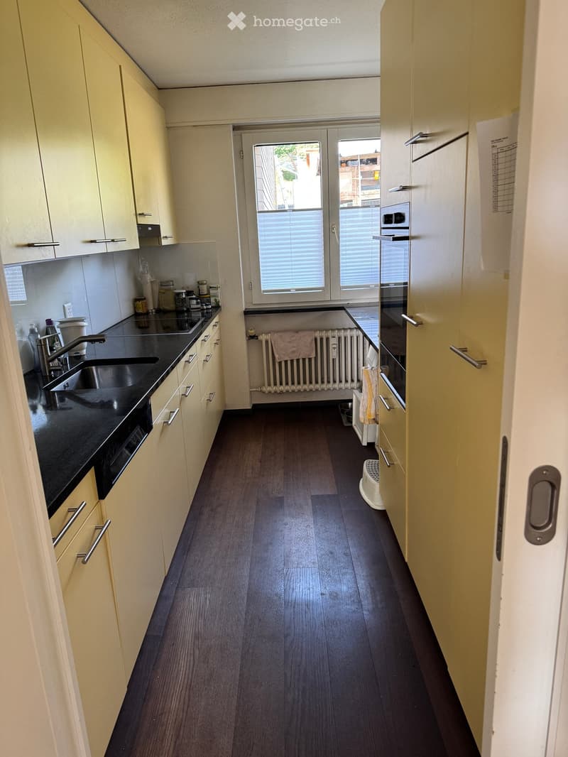 Modern kitchen with light-colored cabinets, dark countertops, and a window with blinds. Includes a sink, faucet, oven, refrigerator, radiator, and towel.