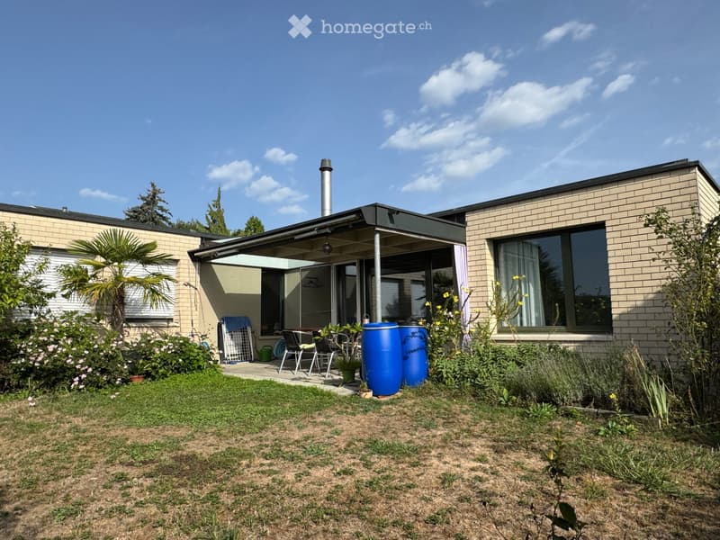 Garden area with a patio, outdoor furniture, and blue barrels, surrounded by plants and trees.