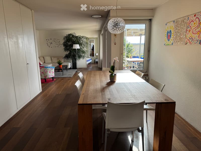 Open-plan living and dining area with wooden flooring, a long dining table with chairs, a sofa, coffee table, floor lamp, and artwork on the walls.