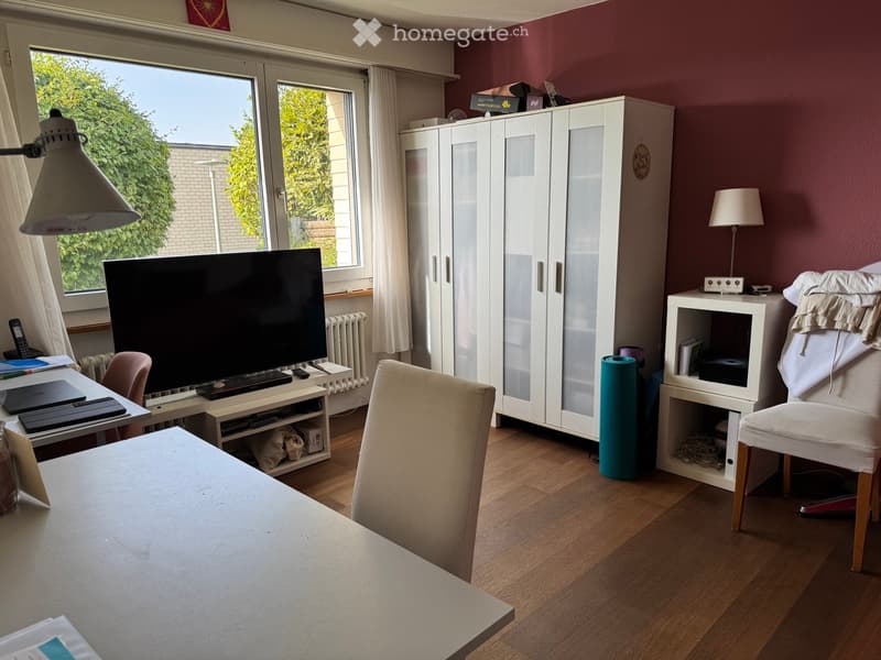 A well-lit room with wooden flooring, a large window with white curtains, a white cabinet, a chair, a table with a laptop, and a screen on a stand.
