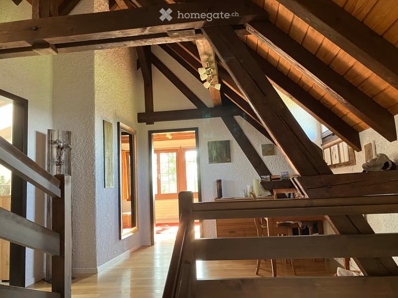 Spacious hallway with wooden beams and exposed wooden ceiling, leading to a room with large windows and a wooden staircase.