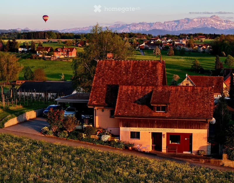 A charming house with a red-tiled roof and a well-maintained garden, situated in a picturesque countryside with mountains in the background.