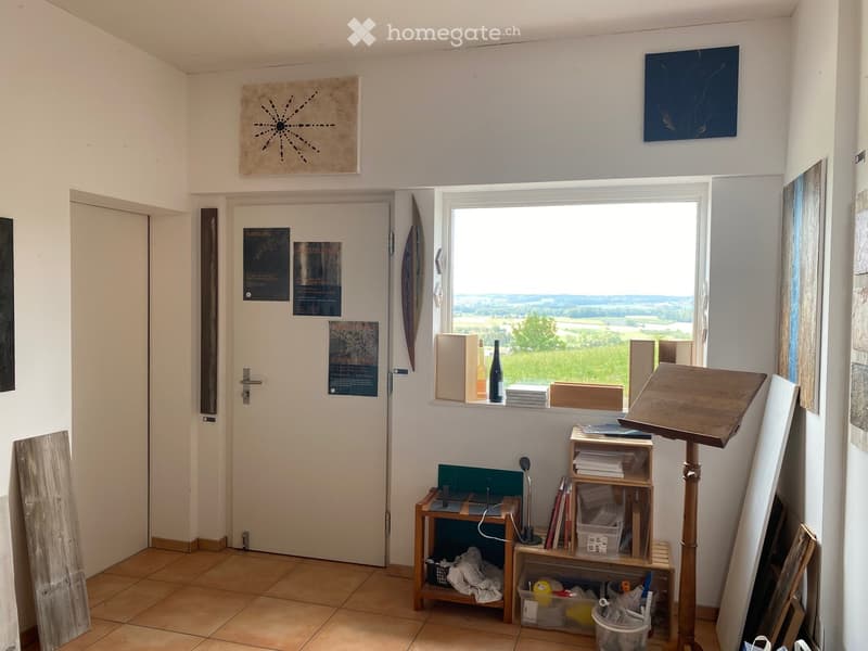 A small, well-lit room with a large window offering a scenic view, featuring a wooden table, shelves, a lamp, a bottle, a stack of books, an easel, and several artworks on the walls.