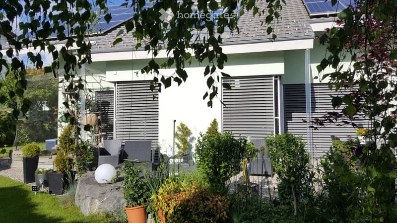 Well-maintained garden with various plants, shrubs, and potted plants, featuring a modern house with solar panels on the roof and a covered patio area with outdoor furniture.