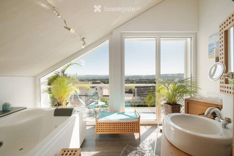 Bright bathroom with large windows offering a scenic view, featuring a bathtub, sink, and a small seating area with a table and chairs.