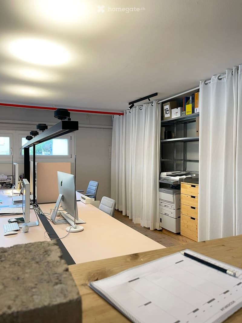 Office space with a large desk, computer setup, chairs, shelves, and a printer. The room has large windows with curtains and a wooden floor.