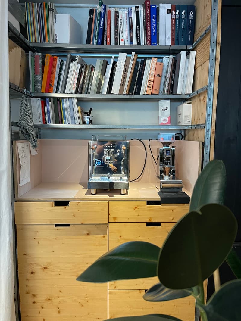 A modern, minimalist kitchen area with a coffee machine, a bookshelf filled with books, and wooden drawers.