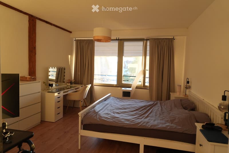 Cozy bedroom with wooden flooring, a single bed with dark bedding, a white desk with a chair, a dresser, and a mirror. Large windows with curtains let in natural light, and a hanging lamp provides additional illumination.