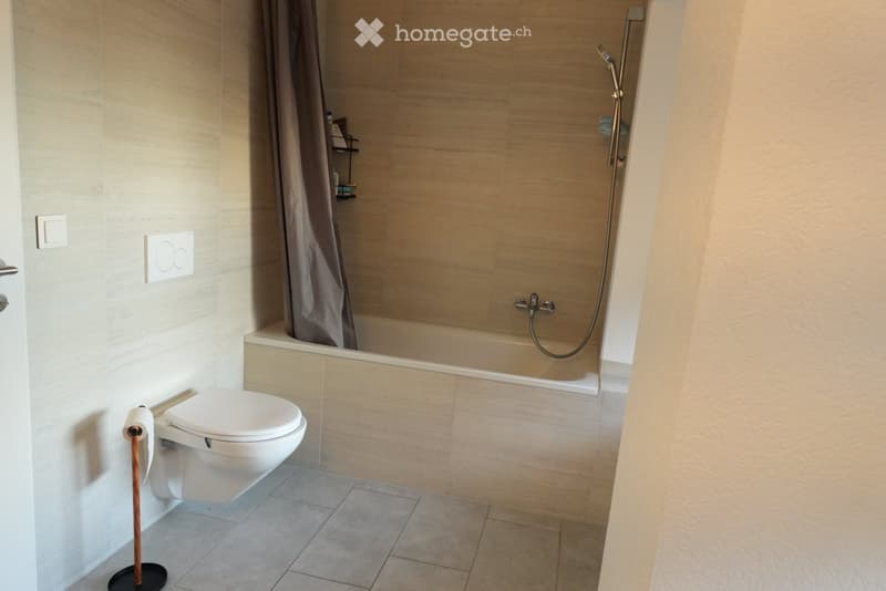 Modern bathroom with a white toilet, a bathtub with a showerhead, and a toilet paper holder. The walls are tiled in a light beige color, and the floor is tiled in a light gray color.