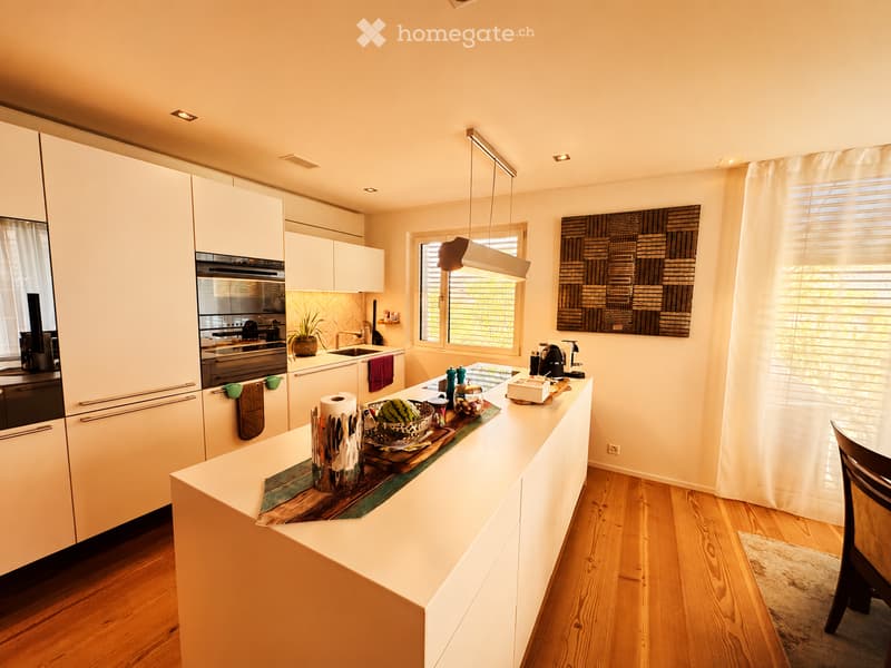 Modern kitchen with white cabinets, a central island, and wooden flooring. Features include a sink, oven, coffee maker, and decorative items on the island.
