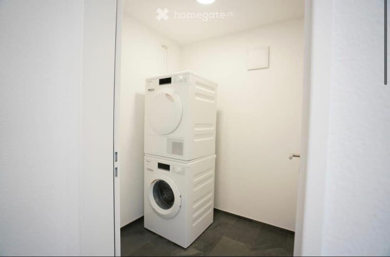 Compact room with a stacked washing machine and dryer unit against a white wall, featuring a dark tiled floor and a closed door.
