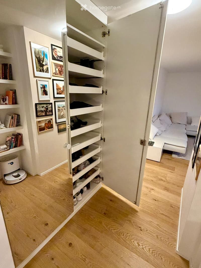 A compact, well-organized room with a bed, shelves filled with books and picture frames, and a robot vacuum on the floor.