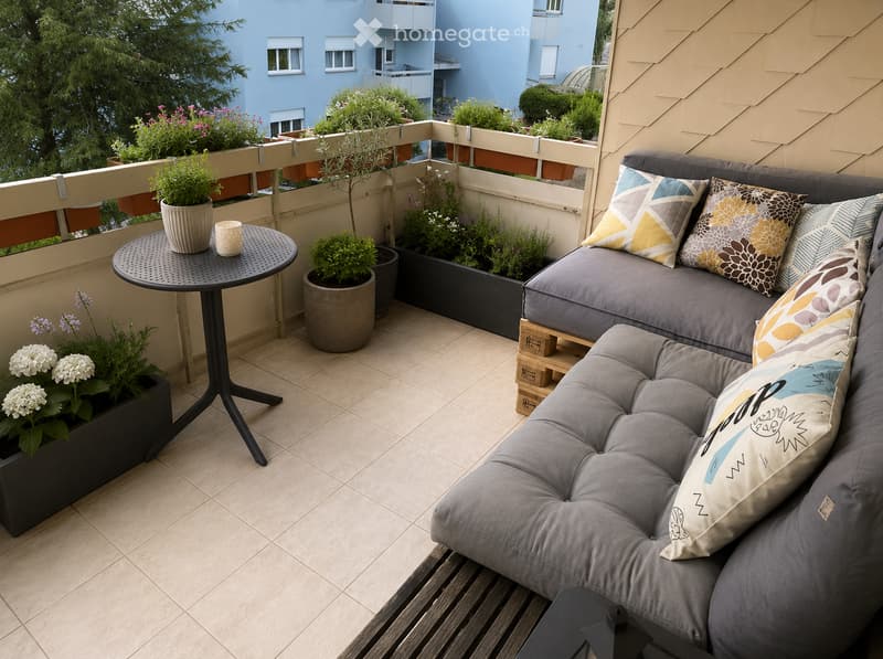 Balcony with a grey sofa, colorful cushions, a small round table with a cup and potted plants, and a view of neighboring buildings.