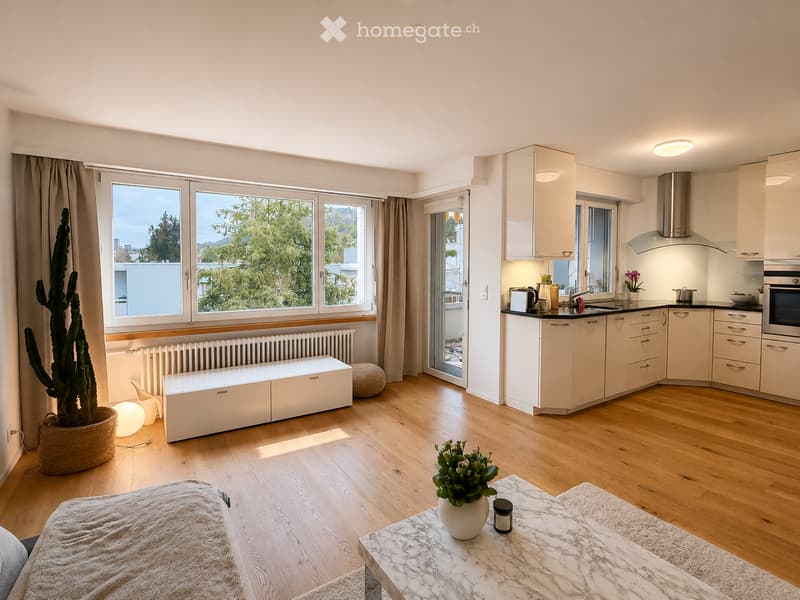 Bright, open-plan living and dining area with a modern kitchen. Features include a white sofa, marble coffee table, potted plants, a tall cactus, and a sleek kitchen with white cabinets, stainless steel appliances, and ample natural light from large windo
