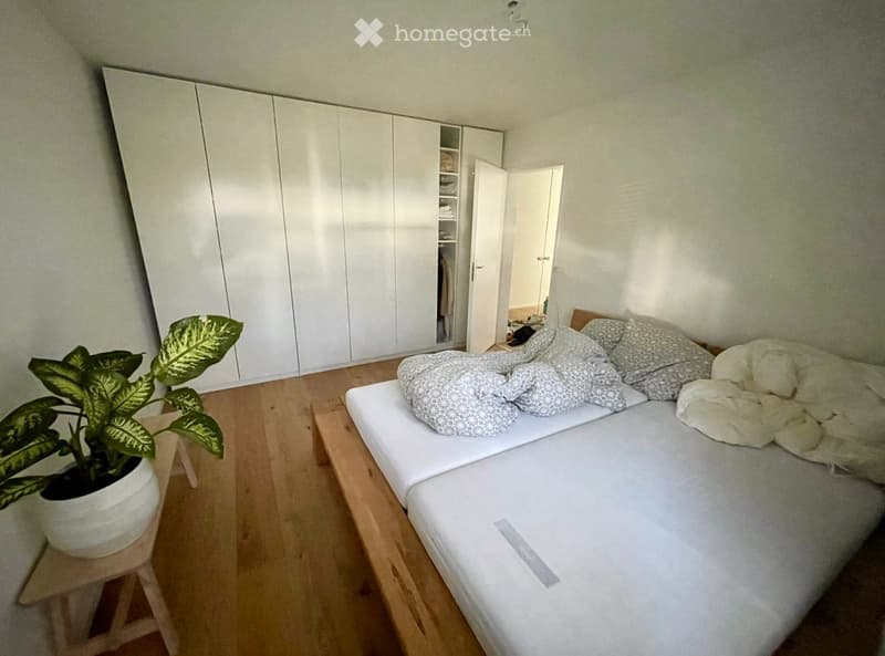 Modern bedroom with a double bed, white wardrobe, a small stool with a potted plant, and a partially open closet revealing more storage space.