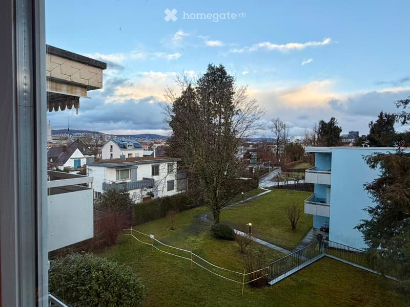 View from a balcony showing a well-maintained garden area with grass, trees, and pathways, surrounded by modern apartment buildings and a cityscape in the background.