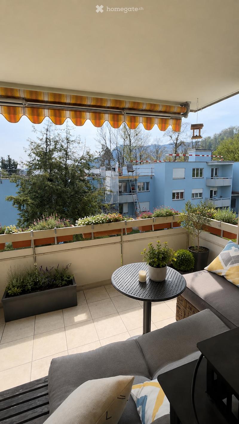Balcony with a grey sectional sofa, small round table, potted plants, and colorful cushions, overlooking a scenic view with trees and buildings.
