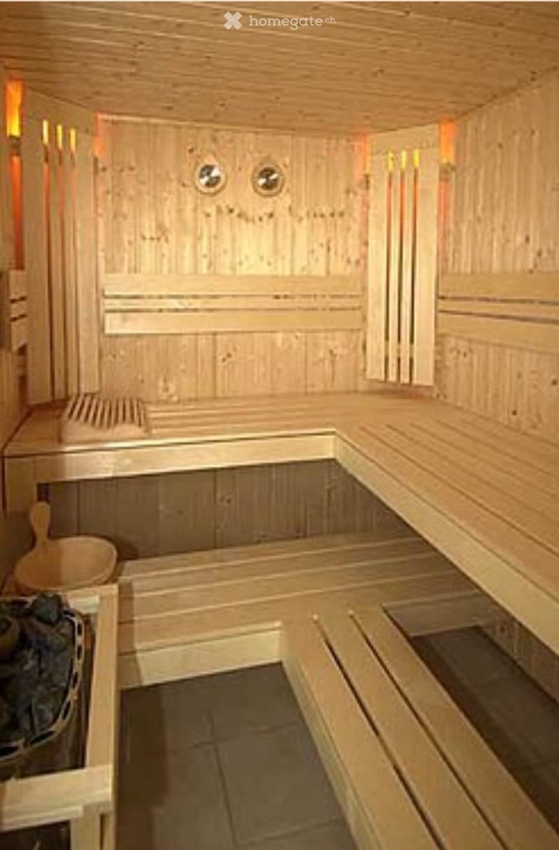 Wooden sauna with benches, wooden walls, ceiling, and floor, featuring a wooden bucket and paddle on the left side.